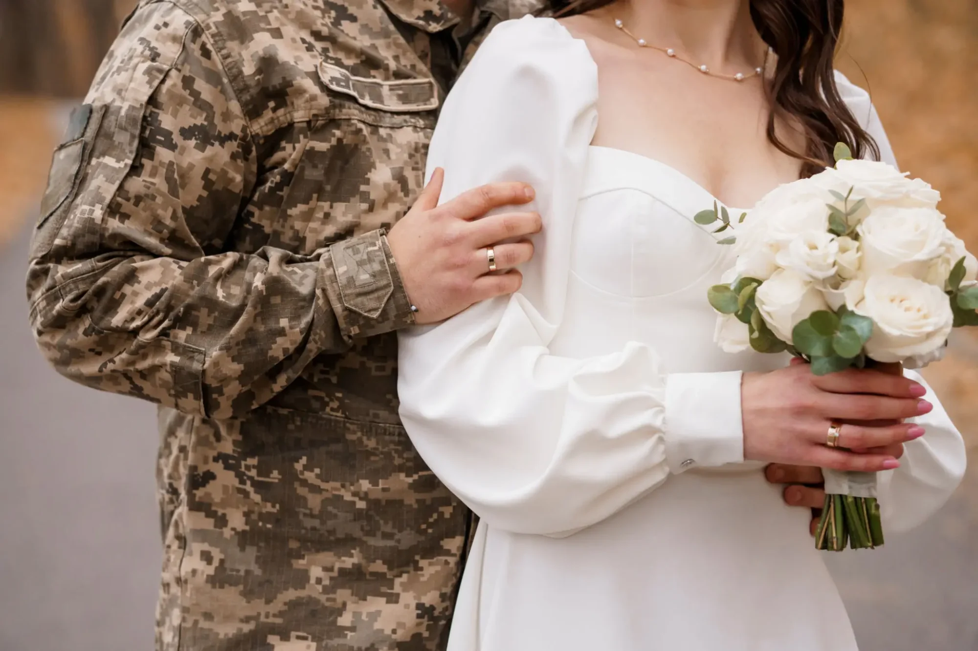 Bride in white dress and groom in camouflage military uniform holding hands.